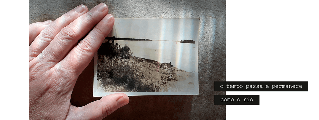 Foto horizontal mostrando parte de uma mão bem iluminada segurando uma foto pequena sobre um fundo bege cheio de sombras. A foto pequena é antiga, sépia, e mostra um grande rio e uma faixa de mato e areia. Na margem mais próxima (que está distante da câmera) vê-se bem ao longe a silhueta de uma canoa e de uma pessoa, que parece estar chegando ou partindo com a canoa. Pequenas faixas verticais de luz que iluminam o fundo bege iluminam também a foto antiga. Há uma frase escrita em duas faixas pretas que começam no lado direito e saem da margem da foto: o tempo passa e permanece / como o rio.