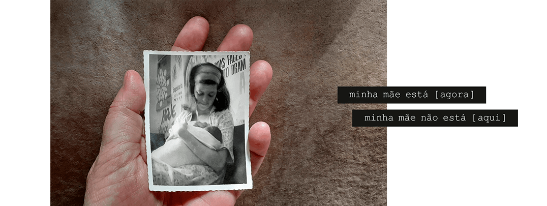 Foto horizontal mostrando no centro uma mão virada para cima, segurando na palma uma foto vertical pequena, antiga, em preto e branco. A mão e a foto estão bem iluminadas e o fundo é bege escuro, com algumas sombras. A foto antiga mostra uma mulher sentada, amamentando um bebê recém-nascido e olhando para ele. Atrás dela estão vários cartazes colados na parede, aparentando ser dos anos 60 ou 70. Há uma frase em duas faixas pretas que começam no lado direito e saem da margem: minha mãe está [ agora ] / minha mãe não está [aqui].