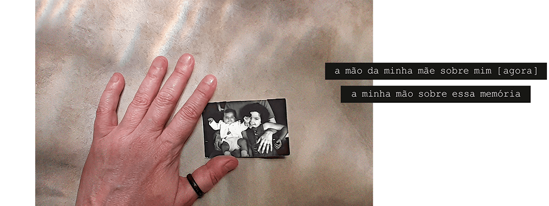 Foto horizontal de uma mão virada para baixo, com os dedos indicador e polegar sobre uma foto pequena, antiga e em preto e branco. Mão e foto estão apoiadas num fundo bege claro. A foto antiga mostra uma criança de três anos e um bebê de dois meses no colo de alguém cuja cabeça não aparece. A mão dessa pessoa se destaca, aberta sobre o corpo da criança maior, e tem um anel escuro num dedo. A mão da foto maior tem um anel escuro no polegar. Há uma frase em duas faixas pretas que começam no lado direito e saem da margem da foto maior: a mão da minha mão sobre mim [ agora ] / a minha mão sobre essa memória.
