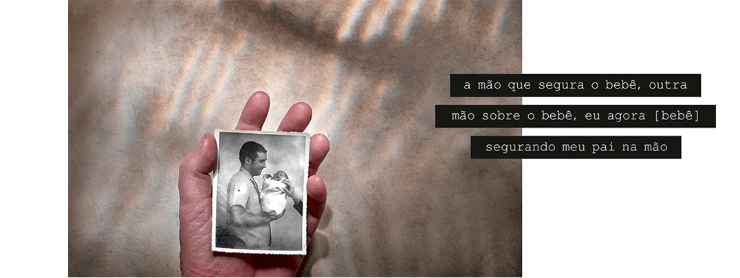 Foto horizontal com fundo bege, muitas luzes e sombras, e linhas verticais de luz vinda de cima. Na parte de baixo, no centro, aparece uma mão voltada para cima, segurando na palma uma pequena foto vertical antiga e em preto e branco. Esta foto mostra um homem de perfil segurando um bebê novinho, todo enrolado num pano branco, e uma outra mão saindo da margem direita e tocando o bebê. Há uma frase sobre três faixas pretas que começam no lado direito e saem da margem: a mão que segura o bebê, outra / mão sobre o bebê, eu agora [bebê] / segurando meu pai na mão.