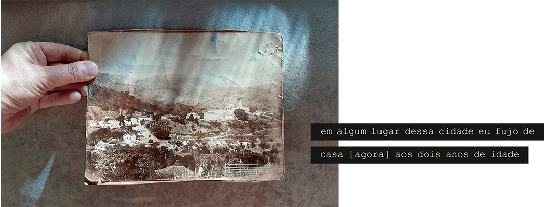 Foto horizontal de uma mão saindo da margem esquerda e segurando uma grande foto antiga, que ocupa quase todo o quadro. O fundo é bege escuro, iluminado por uma luz que vem de cima e forma desenhos na superfície. Esses desenhos também estão sobre a foto antiga, que é sépia e está muito maltratada pelo tempo. Na foto aparece uma pequena cidade vista de um lugar alto, com poucas ruas e muitas árvores, e morros ao longe. Há uma frase escrita em duas faixas pretas que começam na direita saem da margem: em algum lugar dessa cidade eu fujo de casa [ agora ] aos dois anos de idade.