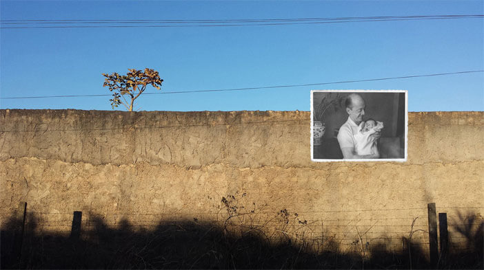 Foto horizontal de uma parede de cimento chapiscado, com uma pequena abertura retangular vertical no lado direito, de onde sai um ramo de folhas verdes. Há sol e sombras escuras sobre a parede. Do lado esquerdo, embaixo de uma sombra grande, está colada uma foto pequena, antiga e em preto e branco. Ela mostra um casal num ambiente rural; o rapaz está meio agachado, meio ajoelhado, de calça e camisa clara. A moça está sentada ao seu lado, com um vestido longo, claro e com estampa miúda.