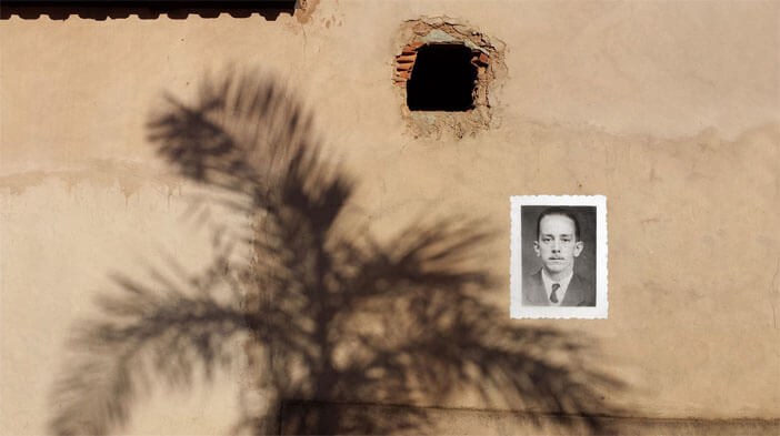 Foto horizontal de uma parede de cimento que foi quebrada para abrir um pequeno buraco quadrado, do tamanho de um tijolo. A parede é marrom-claro e está iluminada pelo sol. Abaixo do buraco preto há uma grande sombra da copa de uma palmeira, e na parte de cima da foto, à esquerda, uma estreita faixa de sombra escura do telhado. Um pouco à direita e abaixo do buraco foi colado um retrato em preto e branco, antigo e com margens brancas, que aparenta ter sido feito para um documento. Ele está sobre uma das folhas da sombra da palmeira e mostra um homem de bigode fino, paletó e gravata.