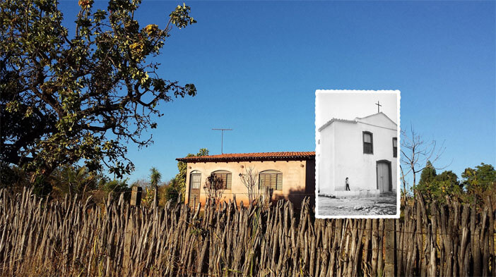 Foto horizontal de uma casinha pequena ao longe, de reboco sem pintura, com uma porta e duas janelas cinzas, também sem pintura. Ela está bem no centro da imagem. À esquerda, mais próxima, há uma grande árvore do cerrado com folhas verdes. Na frente da foto, de fora a fora, há uma cerca rústica feita de galhos secos. O céu é azul e sem nuvens, e a paisagem está iluminada pelo sol do final da tarde. Há uma pequena foto antiga em preto e branco colada sobre a cerca, o lado direito da casinha e o céu. Ela mostra, ao longe, parte de uma capelinha antiga, branca, com uma porta embaixo e duas janelas em cima. Na frente da capelinha há uma criança pequena encostada na parede.