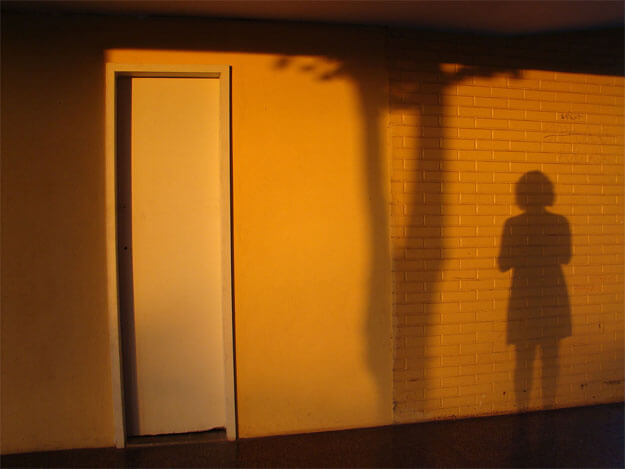 Foto horizontal de uma parede com uma porta estreita e a sombra de uma mulher. A porta é estreita e branca, a parede é bege, mas tudo está amarelo/laranja vivo com o sol do finalzinho da tarde. A porta está à esquerda; do centro para a direita vê-se a sombra de um tronco de árvore e a sombra da mulher com vestido e cabelos curtos, que parece estar tirando a foto. Acima e abaixo há faixas escuras onde o sol não está batendo.