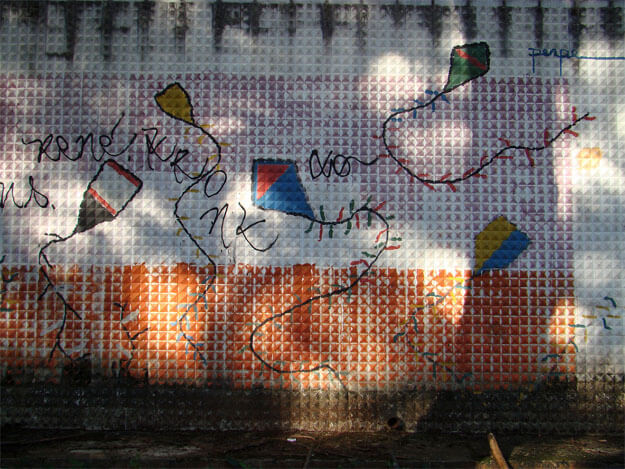 Foto de uma parede com desenhos de cinco pipas e algumas pixações em spray, em harmonia com os desenhos. A parede, vista de frente, está quase toda na sombra, mas tem vivas manchas de luz do sol filtrada por árvores (que não aparecem). As pinturas e pixações estão sobre quatro faixas horizontais feitas, de cima para baixo, por manchas pretas de umidade; pintura rosa; pintura branca; pintura laranja; e outras manchas pretas de umidade. 