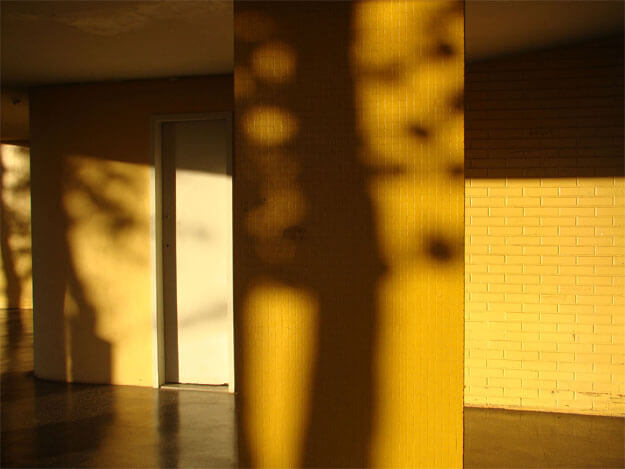 Foto horizontal que mostra vários planos de uma área embaixo de um prédio. Todos os planos estão iluminados pela forte luz amarela do sol poente. Em primeiro plano, ligeiramente à direita, há uma coluna larga, retangular, cobertas de pastilhas amarelas, com a sombra bem escura do tronco de uma árvore. Em segundo plano, alguns metros atrás, está uma parede de tijolinhos amarelos e uma estreita porta branca, com sombras pretas do teto e das árvores (que não aparecem). O chão é de cimento brilhante e também está amarelado, refletindo a luz. O teto é plano e está todo na sombra, mas também recebe o reflexo da luz amarela. Ao fundo, no canto esquerdo, há um último plano, estreito, de uma parede também amarela e com sombra de árvores.(Esta é a última imagem da série.)