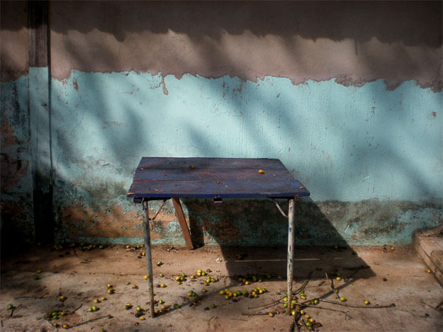 Foto horizontal de uma mesa velha encostada numa parede velha. A mesa está no centro da foto, enquadrada de frente, fazendo uma sombra escura para o lado e para baixo. A parede tem no meio uma faixa de pintura velha, azul claro; por cima, uma faixa de reboco, e por cima do reboco a sombra escura do telhado. Rente ao chão há uma faixa desgastada e manchada de marrom. A mesa tem tampo azul marinho quadrado e três pernas desiguais. O chão é de cimento, com alguns gravetos e muitas frutinhas verdes e amarelas caídas. 