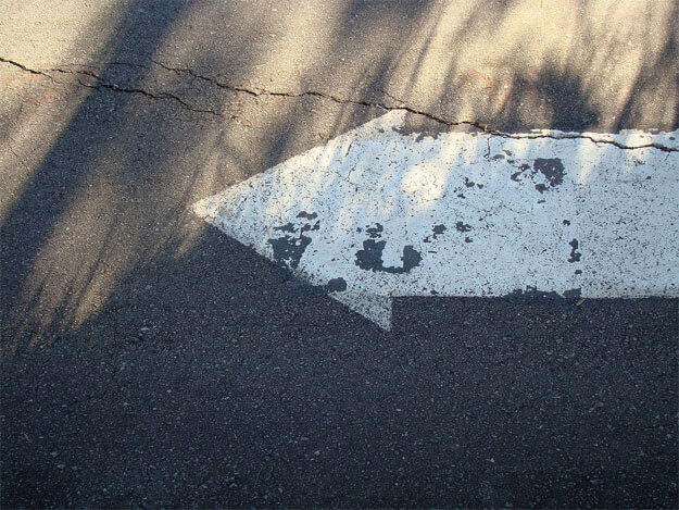 Foto horizontal de uma seta branca pintada no asfalto, apontando para a esquerda. Na parte de cima da foto há sol, e a de baixo está na sombra. Sombras de capim se alongam em direção ao alto da imagem. Na parte ensolarada há uma rachadura horizontal que atravessa toda a foto, descendo ligeiramente e passando por cima da seta, que está quase toda na sombra. 