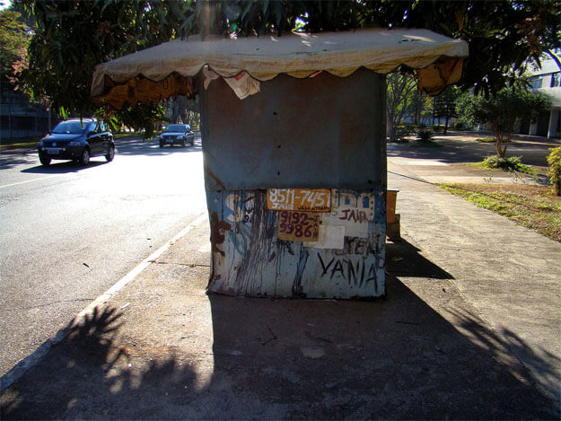 Foto horizontal de uma barraca de rua feita de pedaços de madeirite, com pintura azul velha e desbotada. A barraca está de frente; na parte de baixo lê-se uma palavra grande, que pode ter sido "sapateiro", mas está quase toda coberta por outras coisas escritas: dois números de celular, os nomes Jair e Vânia e alguns papéis ilegíveis. O teto é um velho toldo de lona, com beiral bem comprido, debaixo de uma árvore. A barraca está na calçada na beira de uma rua onde há dois carros passando. O sol vem de trás, de modo que a barraca lança uma grande sombra para a frente, que ocupa toda a parte de baixo da foto.