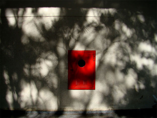 Foto horizontal de uma parede com um retângulo pintado de vermelho vivo, visto de frente, no centro da imagem. Ele é vertical e tem um buraco redondo na parte de cima. A parede é bege claro. Toda a imagem está coberta por sombras que fazem uma espécie de rendilhado quase preto na parede ensolarada; parecem sombras de árvores. Em cima há uma estreita faixa preta onde o sol não bate, e embaixo uma estreita faixa cinza escura de piso.
