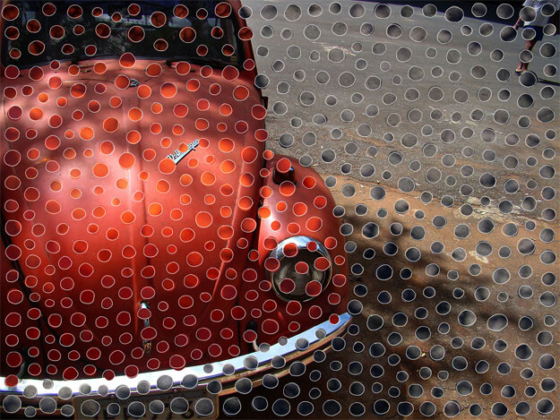 Foto horizontal de um fusca vermelho antigo, do parachoque até o alto do parabrisa dianteiro. Aparece apenas um dos faróis (o outro foi cortado na margem esquerda). Na metade direita da foto vê-se o chão de cimento e asfalto emendados e uma pessoa ao longe, cortada na altura dos ombros, no canto de cima. Há bastante luz do sol e sombras sobre o fusca. Sobre toda a foto foram coladas bolinhas recortadas de outras fotos, em tons de vermelho, vinho, marrom, cinza azulado e cinza chumbo. Estão bem próximas e fazem um degradê acompanhando as cores do fundo. Seu formato é irregular, contornado por uma linha clara.