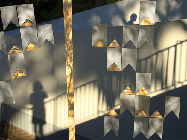 Colagem de uma composição de mastro e bandeirinhas de Volpi sobre uma foto horizontal. As bandeirinhas e o mastro foram recortados de outras fotos (de uma parede branca e outra amarela, com desenhos feitos por sol e sombras). A foto de fundo mostra duas paredes de uma rampa de garagem, com luz do sol e sombras fazendo desenhos. Alguns desses desenhos são sombras de árvores; outros são sombras das paredes, retas, que criam a ilusão de diversos planos inclinados. No alto, à direita, aparece a sombra de uma pessoa de perfil, dos ombros para cima. No canto de baixo, à esquerda, há a sombra de uma menina e de uma grade baixa, que sobe em direção à direita.