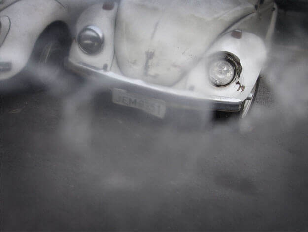 Foto horizontal mostrando a parte de baixo de um fusca branco, bastante enferrujado. Ele é visto de frente, no centro, cortado pela margem de cima. Há outro fusca branco colado a ele, cortado pela margem esquerda; vê-se apenas um pneu e parte do parachoque. Na metade de baixo da foto vê-se o chão escuro. Toda a imagem está desfocada pelo anteparo, menos na área do farol à direita. O enquadramento pende ligeiramente para a direita.