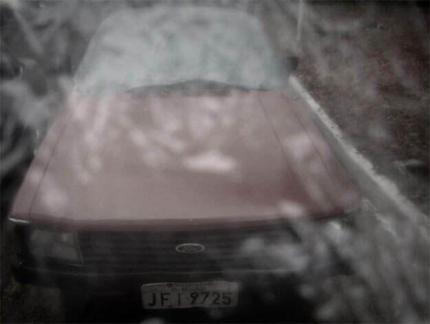 Foto horizontal de um carro vermelho desbotado e amarronzado pela baixa saturação. Ele aparenta ser dos anos 80, com design em linhas retas. Está enquadrado bem de perto e de frente, da placa ao topo, em perspectiva. Junto ao carro, no lado direito, há um meio-fio branco e o que parece ser grama seca ou terra. Toda a imagem está desfocada pelo anteparo, e boa parte está coberta com manchas brancas.