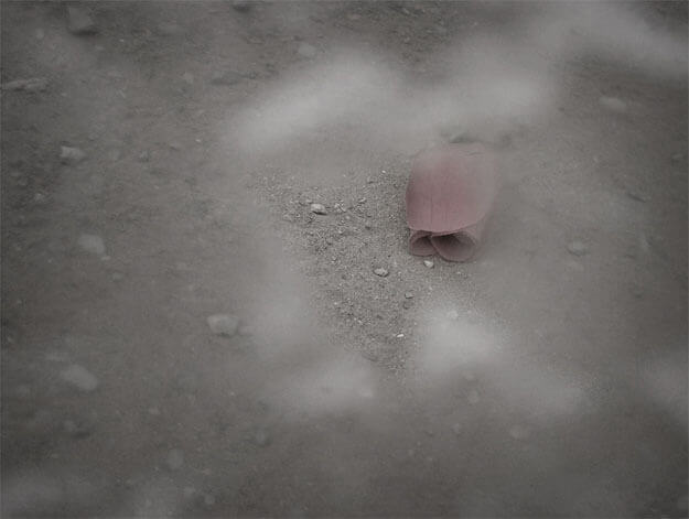 Foto horizontal mostrando uma rosa vermelha de tecido, com a cor esmaecida pela baixa saturação. Ela é pequena e aparece do centro para o canto superior direito. Está sobre um chão de terra com pequenas pedrinhas, que forma todo o fundo da imagem. Tudo está desfocado pelo anteparo, menos uma área onde está o topo da rosa, virado para baixo, e uma parte do chão.