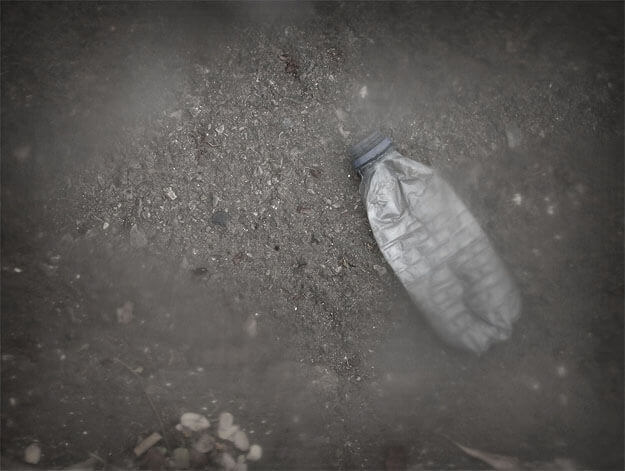Foto horizontal de uma garrafa de água mineral amassada até ficar quase plana. Está sobre um chão de cimento com algumas pedrinhas. A garrafa está inclinada para a esquerda, a meia altura, no lado direito da imagem. Há uma grande área central que não está desfocada pelo anteparo, onde se vê melhor o chão e a parte de cima da garrafa. A foto é bastante escura, e a garrafa se destaca porque reflete a luz.