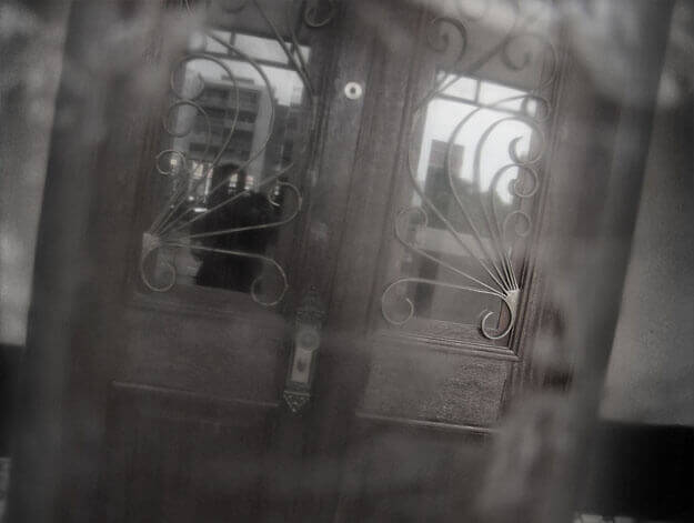 Foto horizontal com o detalhe de uma porta de madeira bem conservada, com dois retângulos de vidro refletindo prédios ao fundo. Sobre o vidro há uma grade de ferro trabalhado com desenhos curvos e harmônicos. O enquadramento está inclinado, de modo que a porta parede pender para a direita. Vê-se uma pequena faixa de parede dos lados. Toda a imagem está desfocada pelo anteparo na lente, exceto por uma área muito pequena onde se vê uma das curvas da grade.