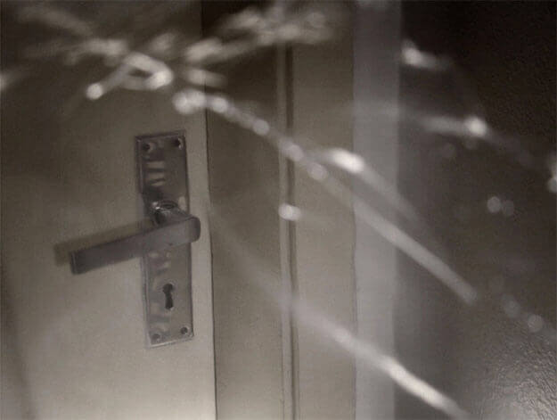 Foto horizontal da maçaneta barata de uma porta comum, branca, fechada, e um pedaço da parede. A foto é bastante escura. O anteparo na lente faz vários traços desfocados e brancos sobre quase toda a imagem. Os traços lembram rachaduras.