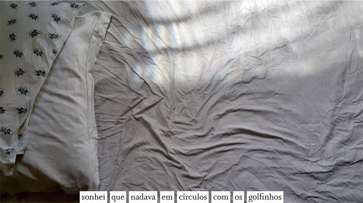 Foto horizontal colorida mostrando um lençol amarrotado e parte de dois travesseiros, um sobre o outro, no lado esquerdo. O travesseiro de cima tem estampa de pequenas flores em azul escuro sobre fundo branco; o outro travesseiro e o lençol são brancos. Três faixas de luz do sol filtrada pela janela ocupam a parte de cima da foto. Colada à margem de baixo há uma frase sobre uma faixa branca: sonhei que nadava em círculos com os golfinhos. As faixas de luz e as dobras do lençol são ligeiramente curvas, lembrando ondas.