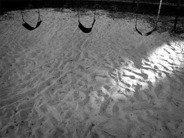 Três balanços de parque infantil aparecem na parte de cima, quase saindo do quadro. Estão pendurados por correntes e são curvos, talvez de couro. Na parte de baixo, que ocupa quase toda a foto, está o chão de areia branca e fofa, todo ondulado por pisadas. Uma pequena faixa de luz do sol entra pelo lado direito e desce até o centro da foto, iluminando também a corrente do último balanço.