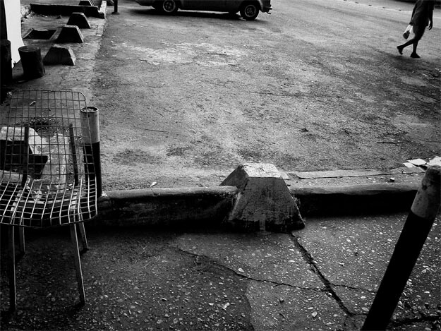 Foto de... nada. Composição estranha mostrando o chão de um estacionamento asfaltado e a silhueta escura de uma pessoa ao longe, do ombro para baixo, no canto direito. No centro, a silhueta da parte de baixo de um carro, só do meio das rodas para baixo. À direita, um meio fio, um bueiro aberto ao longe, um toco de árvore e cinco saliências de concreto enfileiradas, em forma de tronco de pirâmide, se aproximando até sair do quadro. Embaixo, uma cadeira de ferro cortada pela margem esquerda, de trama quadriculada, sem a almofada. Ao longo da parte de baixo, o meio fio com outra saliência de concreto, a calçada de cimento e um poste de metal preto, saindo do quadro na margem direita.