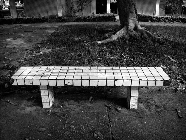 Banco de cimento revestido de pequenos azulejos brancos e quadrados, faltando alguns pedaços. Ele está enquadrado de frente e centralizado na parte de baixo da foto. Na sua frente, uma calçada de cimento. Atrás há um gramado, o tronco de uma árvore com grossas raízes, uma calçada ao fundo e a parte de baixo de um prédio, revestido com os mesmos azulejos brancos e velhos. Do lado esquerdo, uma faixa cimentada desce até a calçada em frente ao banco. 