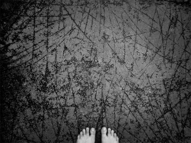 Detalhe de um chão de cimento todo manchado e arranhado, como se tivesse sido riscado em todas as direções antes do cimento secar totalmente. Esses arranhões são bem escuros, assim como as manchas entre eles. Na parte de baixo da foto, bem no centro, aparece a ponta de dois pés descalços, muito brancos e sujos. 