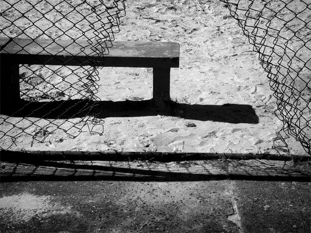 Detalhe de um banco de cimento num parque infantil, com chão de areia branca e uma calçada na parte de baixo. No limite entre a calçada e a areia há uma cerca de alambrado com um grande buraco, mais ou menos no centro da foto. Ele emoldura o lado direito do banco. O banco está colado à margem esquerda da foto e lança uma sombra escura na areia.