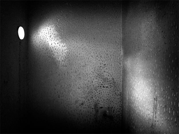 Foto de um aposento vazio, muito pequeno e escuro, muito apertado. Vê-se a parede do fundo e as laterais. Na parede da esquerda, a mais estreita e mais escura, há uma pequena abertura redonda por onde entra um pouco de luz. As paredes estão manchadas e inteiramente cobertas por pequenos buracos feitos manualmente.