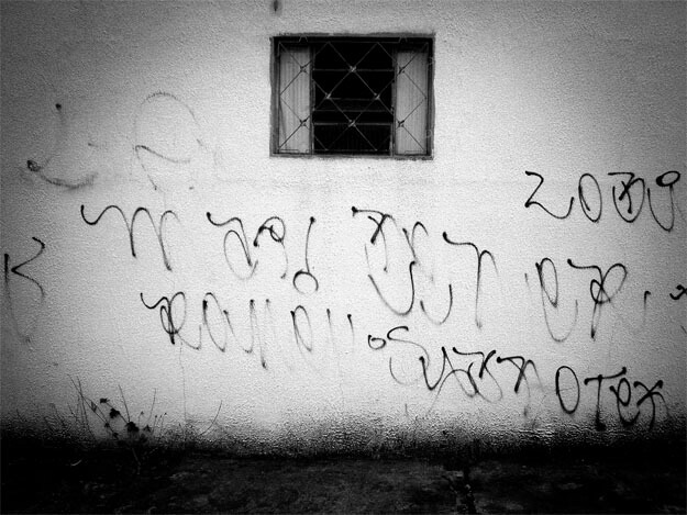 Na parte de cima, no meio da foto, há uma pequena janela basculante retangular, protegida por uma grade. O restante do quadro é uma parede branca, suja e cheia de pixações. Há uma pequena faixa de calçada embaixo, quase preta, manchada e com alguns ramos de mato crescendo. 