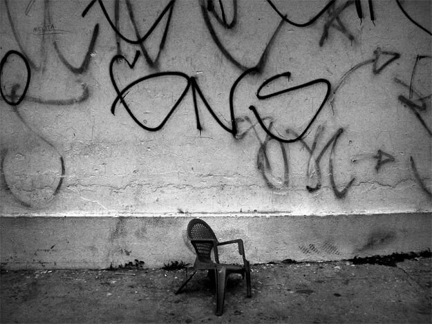 Foto de uma cadeira de plástico de jardim, infantil, escura e com uma perna quebrada, enconstada em uma parede branca, suja e manchada. Ela está sobre o chão de cimento, na parte mais baixa da foto, no centro. Acima da cadeirinha, na parede que ocupa a maior parte da imagem, há dezenas de pixações em cores escuras.