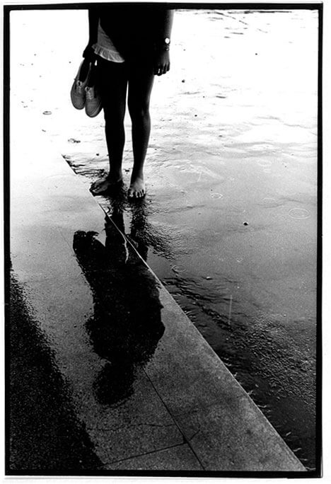 Foto vertical de parte do corpo de uma jovem e de seu reflexo no chão molhado. Ela está escura, na contraluz, e aparece só da cintura para baixo. Está descalça, com os braços junto ao corpo; segura um par de tênis na mão e usa short branco e blusa preta. O fundo está branco na parte de cima, e os tons vão escurecendo em direção à margem inferior. Há uma linha atravessando a foto em diagonal, do canto esquerdo ao direito. Ela delimita o asfalto e um piso liso, onde aparece o reflexo da jovem, de cabeça para baixo. Todo o chão está molhado e vê-se pequenos círculos onde as gotas de chuva tocam o asfalto.