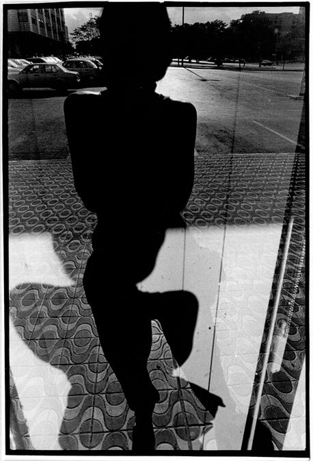 Foto vertical em preto e branco com o reflexo de uma pessoa num vidro espelhado. É uma silhueta preta, cortada no alto da cabeça e na ponta dos pés pelas margens da foto. Está com os braços recolhidos junto ao corpo e as pernas ligeiramente afastadas. Vê-se uma calçada atrás dela, até a altura dos braços, com desenhos simétricos de ondas e círculos. Acima há um estacionamento asfaltado, alguns carros, parte de um prédio e árvores ao longe. O brilho do sol se reflete no asfalto, clareando a área atrás da pessoa. Na metade de baixo da foto há um desenho intrigante sobre a calçada, curvo e distorcido; parece ser a sombra da pessoa sobre o vidro espelhado.