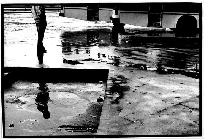 Foto horizontal de um homem numa rodoviária. Ele está a meia distância, à esquerda, cortado na altura dos ombros pela margem de cima. Vê-se dois ônibus estacionados mais adiante, à direita, cortados acima das rodas. O homem está numa pose tranquila, de pé, com as mãos no bolso, parado, virado para a direção dos ônibus. Numa poça d'água abaixo dele aparece o seu reflexo por inteiro. Todo o piso está molhado. O homem está na contraluz, bem escuro, com a silhueta recortada pela luz que entra do lado esquerdo. 