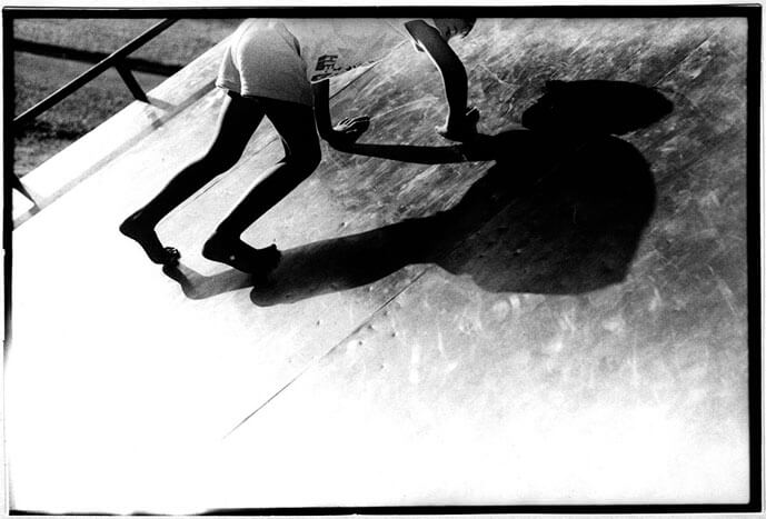 Foto horizontal da sombra de uma criança pequena sobre uma superfície metálica, inclinada em 45 graus. A criança é magrela e está de short e camiseta, subindo pela superfície metálica, apoiando-se nos pés e nas mãos. Sua imagem está cortada no alto das costas e na maior parte da cabeça. A sombra está inteira, bem definida e bem maior que a criança. Um excesso de luz invade a margem esquerda e a parte de baixo da foto, deixando essas áreas totalmente brancas.