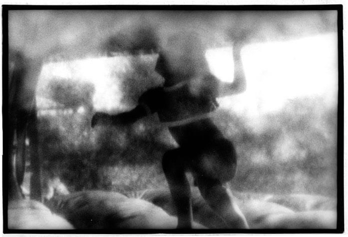 Foto horizontal de uma criança pequena dentro de um pula-pula inflável, vista de fora. A parede de plástico transparente do pula-pula deixa toda a imagem desfocada, cheia de borrões. A menina aparece dos pés para cima; tem a cabeça virada para cima, um dos braços à frente do corpo e o outro atrás, dobrado para cima em ângulo reto. Está no meio de um pulo, com a perna da frente levantada no mesmo ângulo ângulo do braço.