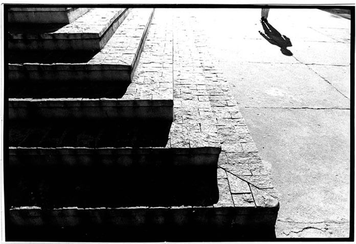 Foto horizontal mostrando os pés de um homem que anda em direção à câmera, no canto de cima, à direita. A sombra de todo o seu corpo se alonga à sua frente. Há uma escadaria em L fazendo uma linha diagonal que atravessa a foto de alto a baixo, da esquerda para a direita. Acima dessa diagonal o chão está claro, quase branco; abaixo, está quase preto por causa da sombra dos degraus. Na imagem vê-se apenas seis degraus da escada, a larga calçada de cimento e a sombra do homem.