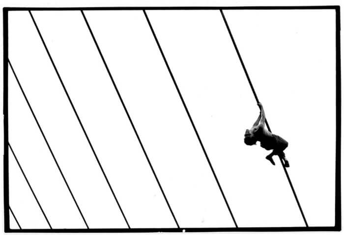 Foto horizontal com fundo todo branco, atravessado por sete fios pretos em diagonal. Os fios se distanciam para a esquerda, em perspectiva. No primeiro, junto à margem direita, há um rapaz pendurado pelas mãos e pernas, de calça, sem camisa, a meia distância. É quase uma silhueta, iluminada pela luz que vem de cima. Seu corpo está estendido numa pose graciosa, com as pernas cruzadas, os braços esticados e o pescoço arqueado para trás. O rosto está voltado para a câmera, de cabeça para baixo, e ele sorri. (Esta é a última foto da série.) 