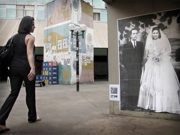 Foto horizontal no mesmo local anterior. A parede com a foto antiga ocupa um terço da imagem, à direita. No canto esquerdo há uma mulher passando, de lado, olhando para a foto com ar sério, quase carrancudo. Tem cabelos lisos na altura do ombro e está de blusa e calça preta.
