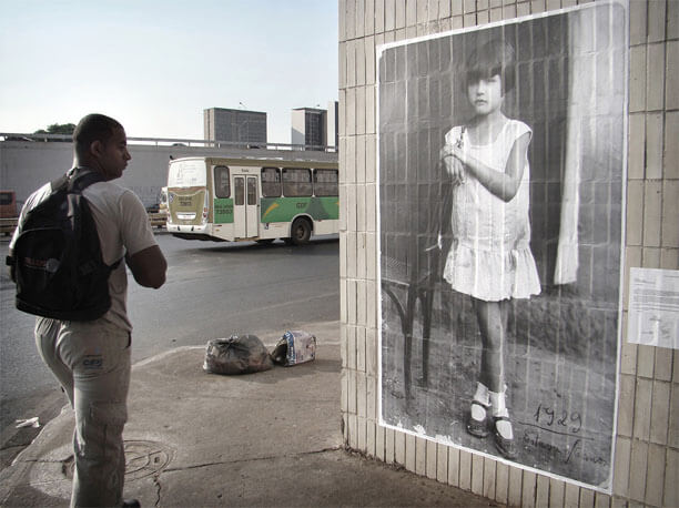 Foto horizontal de um homem passando e olhando para a foto antiga de uma menina pequena, que ocupa a metade direita da imagem. O homem está no lado esquerdo, em meio perfil, sério. Vê-se a calçada, dois sacos de lixo, um ônibus passando na rua e, no fundo, a parte de cima de três prédios que surgem detrás de um paredão com guarda-corpo. (Quem é de Brasília vê que o homem está saindo da rodoviária do Plano Piloto.) Na foto antiga a menina aparece em tamanho um pouco maior que o natural. Ela está de pé num chão de terra batida, junto a uma cadeira de madeira. Tem cabelos curtos e lisos, com corte chanel. Usa um vestido branco curto, de cintura baixa, sapatos boneca bem usados e meias brancas curtas. Suas pernas estão sujas. Ela apoia um dos braços no encosto da cadeira e suas mãos estão juntas. Está ligeiramente de perfil, com o olhar ao longe, e tem as pernas cruzadas, numa pose harmônica e claramente arranjada pelo fotógrafo. No cantinho da foto está escrito "1929", e, embaixo, "Estava c/ 6 anos".