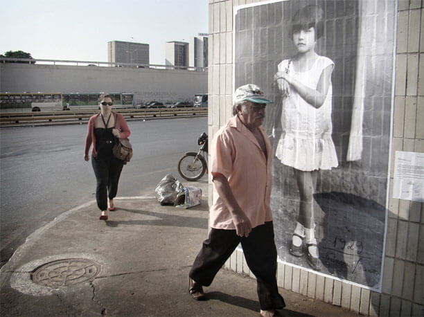 Foto horizontal no mesmo local da imagem anterior. Em frente à foto antiga da criança está passando um senhor de boné, de cabelos e bigode brancos, em meio perfil, apressado e sério, olhando para o chão à frente. Alguns metros atrás dele vem andando uma mulher jovem, de óculos escuros, também séria e olhando para a frente. A pista atrás dela está vazia; na pista mais ao longe há muitos carros e ônibus.