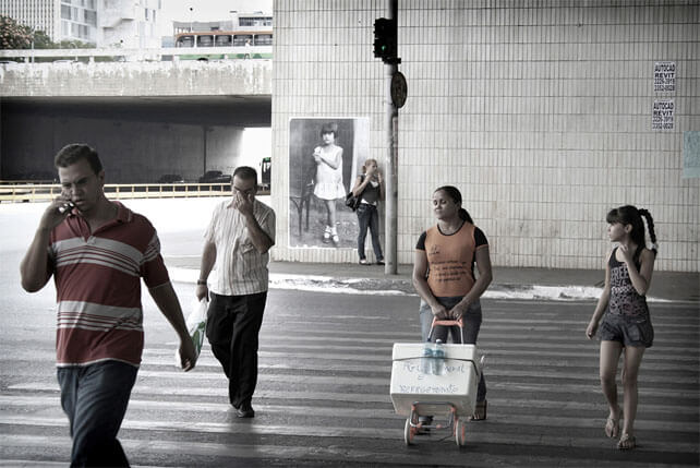 Foto horizontal do mesmo local da imagem anterior, agora enquadrado de frente e de longe, do outro lado da rua. Quatro pessoas atravessam a faixa de segurança, de frente para a câmera: um homem jovem falando ao celular; um homem de cabeça baixa, apertando os olhos com a mão; uma vendedora de água, empurrando um isopor num carrinho; e uma menina de 10 ou 11 anos. Do outro lado da rua vê-se o paredão onde está colada a foto antiga, no canto esquerdo da parede e no centro da foto maior. Encostada à foto antiga há uma mulher jovem, em meio perfil, e à sua frente um poste com um semáforo. Ao fundo vê-se um viaduto, ônibus, carros e prédios.