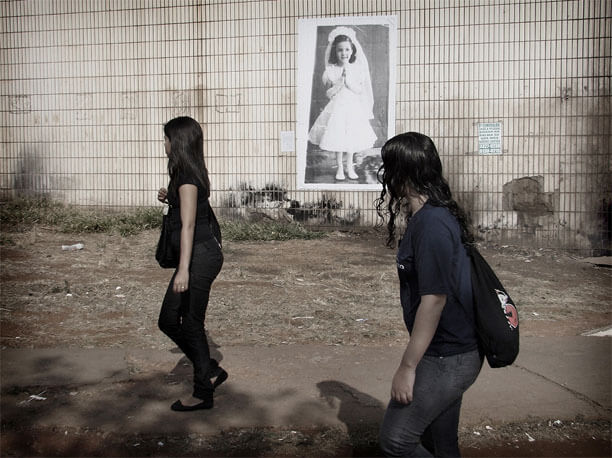 Foto horizontal de duas jovens passando de perfil, com os rostos ocultos, da direita para a esquerda. Elas têm cabelo comprido e escuro e usam roupas escuras; estão afastadas uma da outra. Vê-se uma calçada de cimento e depois o chão de terra com mato seco aparado. Da metade da foto para cima, enquadrado de frente, há um paredão revestido de ladrilhos claros, velhos e sujos. A parede tem grandes manchas escuras.Entre as duas moças vê-se uma foto vertical colada na parede, a cerca de oito ou nove metros de distância da câmera, mostrando uma menina de cabelos escuros, com seis ou sete anos, sorrindo e olhando ligeiramente para o lado. Está de mãos postas e toda de branço: vestido, véu e grinalda, luvas, meias e sapatos.