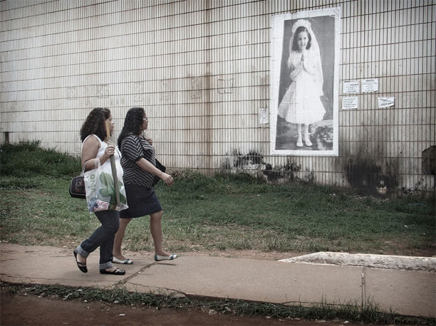 Foto horizontal de duas mulheres passando juntas na calçada, da esquerda para a direita, de perfil, olhando para a foto da menina. A foto antiga está enquadrada em perspectiva e mais de perto que na imagem anterior.