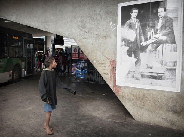 Foto horizontal de um menino de uns 11 anos, descalço, de short e casaco de moletom; as roupas são tão grandes para ele que as mangas cobrem totalmente suas mãos. Ele está em meio perfil, em movimento, tem a boca entreaberta e olha para cima e para a direita, em direção à foto antiga colada na parede. Atrás do menino, bem mais longe e numa área escura da imagem, há um ônibus estacionado e várias pessoas passando. A foto antiga é vertical e está sobre uma parede bem iluminada, de concreto e com formato incomum. A parede sai da parte de cima da imagem, à esquerda, e desce até quase a parte de baixo, à direita. A foto na parede mostra dois homens, um de meia idade e um mais jovem, virados em meio perfil, um para a esquerda e outro para a direita. Ambos estão sérios e olham ao longe. O mais velho, à direita, está sentado sobre a grade de um carrinho industrial de movimentação de mercadorias, com as pernas cruzadas e um dos pés no piso do carrinho. Abaixo vê-se um pequeno rodízio e o chão de terra batida. O homem está de paletó escuro, calça clara e gravata borboleta, e segura um chapéu panamá em uma das mãos. O rapaz ao seu lado está de pé e segura um chapéu panamá debaixo do braço. Ele veste terno escuro e gravata comum. Parte do lado esquerdo da foto original, embaixo, foi desgastada pelo tempo até ficar branca, e há pequenas manchas sobre o resto da superfície.