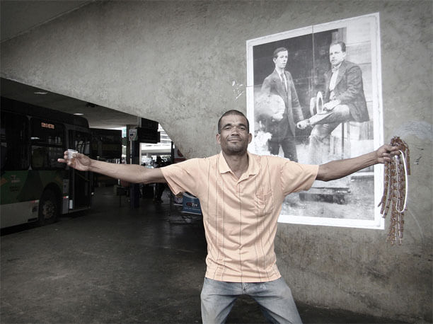 Foto horizontal no mesmo local da foto anterior, mostrando em primeiro plano um homem de braços bem abertos, olhando para a câmera com um meio sorriso e um olhar gaiato. Na mão esquerda ele segura uma dúzia de fileiras de saquinhos de mel, desses que se vende para chupar em porções individuais. Na mão direita segura um copinho plástico com algo que pode ser mel ou cachaça amarela. Seu ombro e braço direitos estão na frente da foto colada na parede, bem visível atrás dele. Ao fundo, na área mais escura, vê-se um ônibus estacionado e pessoas na rodoviária. 