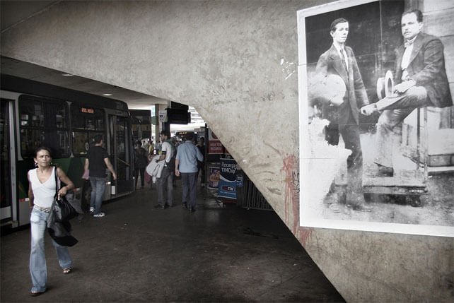 Foto horizontal no mesmo local da anterior, agora enquadrando mais de perto a foto antiga, que está encostada à margem direita e à de cima. No lado esquerdo, bem na ponta, está uma mulher andando apressada em direção à câmera e olhando de lado para a foto antiga, com a testa franzida. A mulher está bem iluminada. Atrás, na área mais escura, vê-se um ônibus estacionado e outras pessoas a poucos metros; ao fundo vê-se a rodoviária com pessoas circulando.