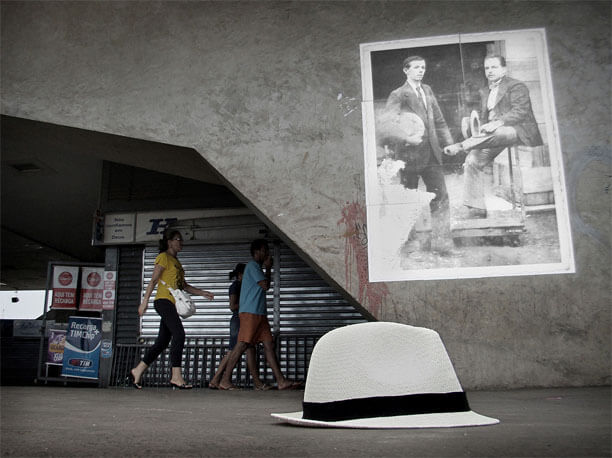 Foto horizontal no mesmo local, mostrando em primeiro plano, no chão, um chapéu de palha com a aba curta e uma faixa preta acima da aba. A foto foi enquadrada do chão, mostrando o chapéu bem de perto. Acima e um pouco à direita dele vê-se a foto antiga na parede. Atrás da parede há um quiosque com parede de metal, alguns cartazes com propagandas e três pessoas passando de perfil, da esquerda para a direita, olhando em frente.