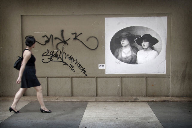 Foto horizontal mostrando uma parede marrom acinzentado com uma grande foto antiga do lado direito. A parede está enquadrada de frente. No lado esquerdo há uma mulher de perfil andando em direção à direita, de cabelo preto e liso até a altura das orelhas. Ela está de vestido curto e preto, sapato preto de salto baixo e bolsa preta. Entre a mulher e a foto antiga há três nomes pixados na parede, com spray preto. A foto é oval, está sobre fundo branco e parece datar do final dos anos 1800 ou começo dos 1900. Mostra duas mulheres do peito para cima, em meio perfil, sérias, olhando ao longe, com chapéus de abas largas. As duas parecem olhar na direção da mulher que preto, que está olhando para elas.